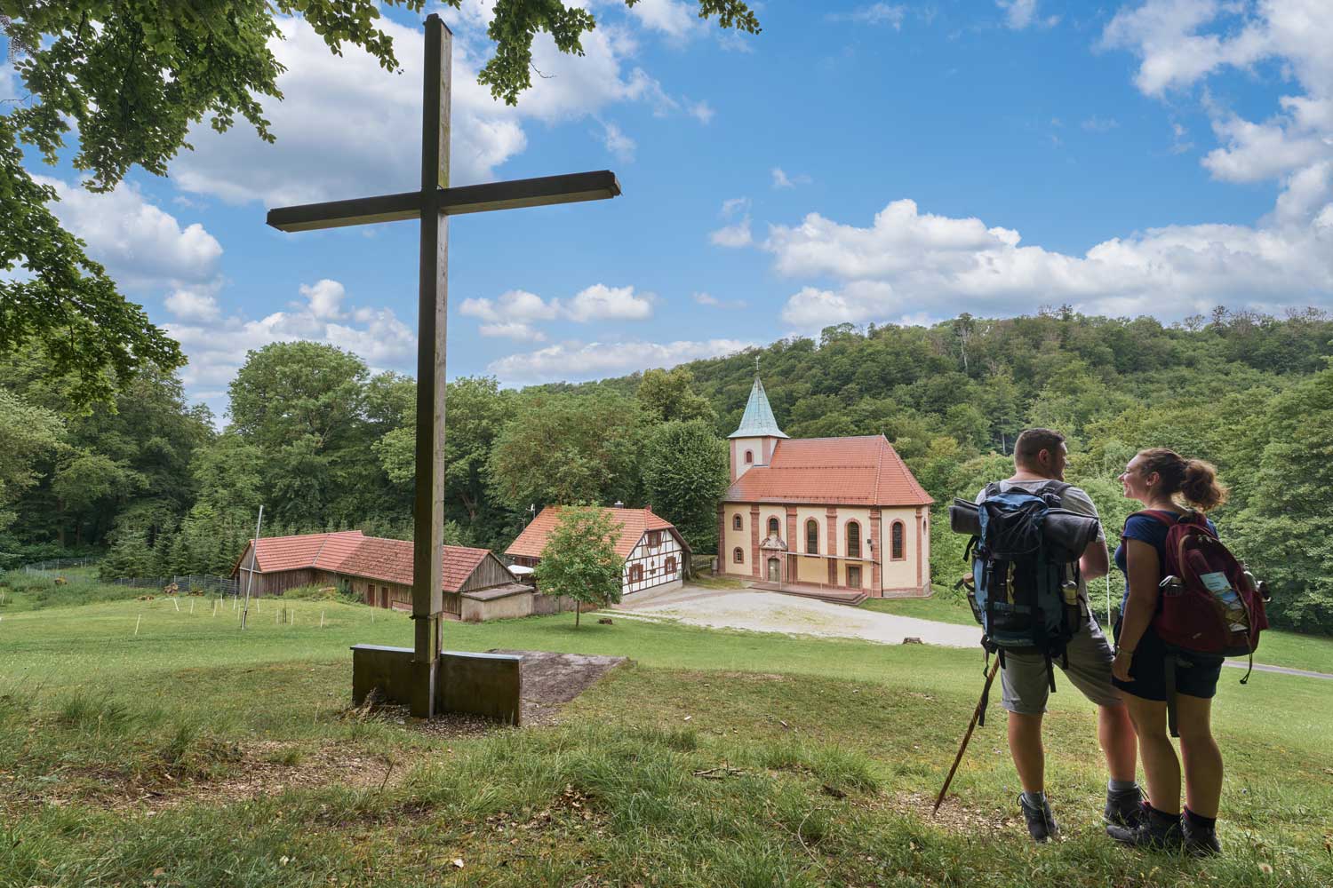 Zwei Wandernde neben einem Gipfelkreuz schauen auf eine Kirche